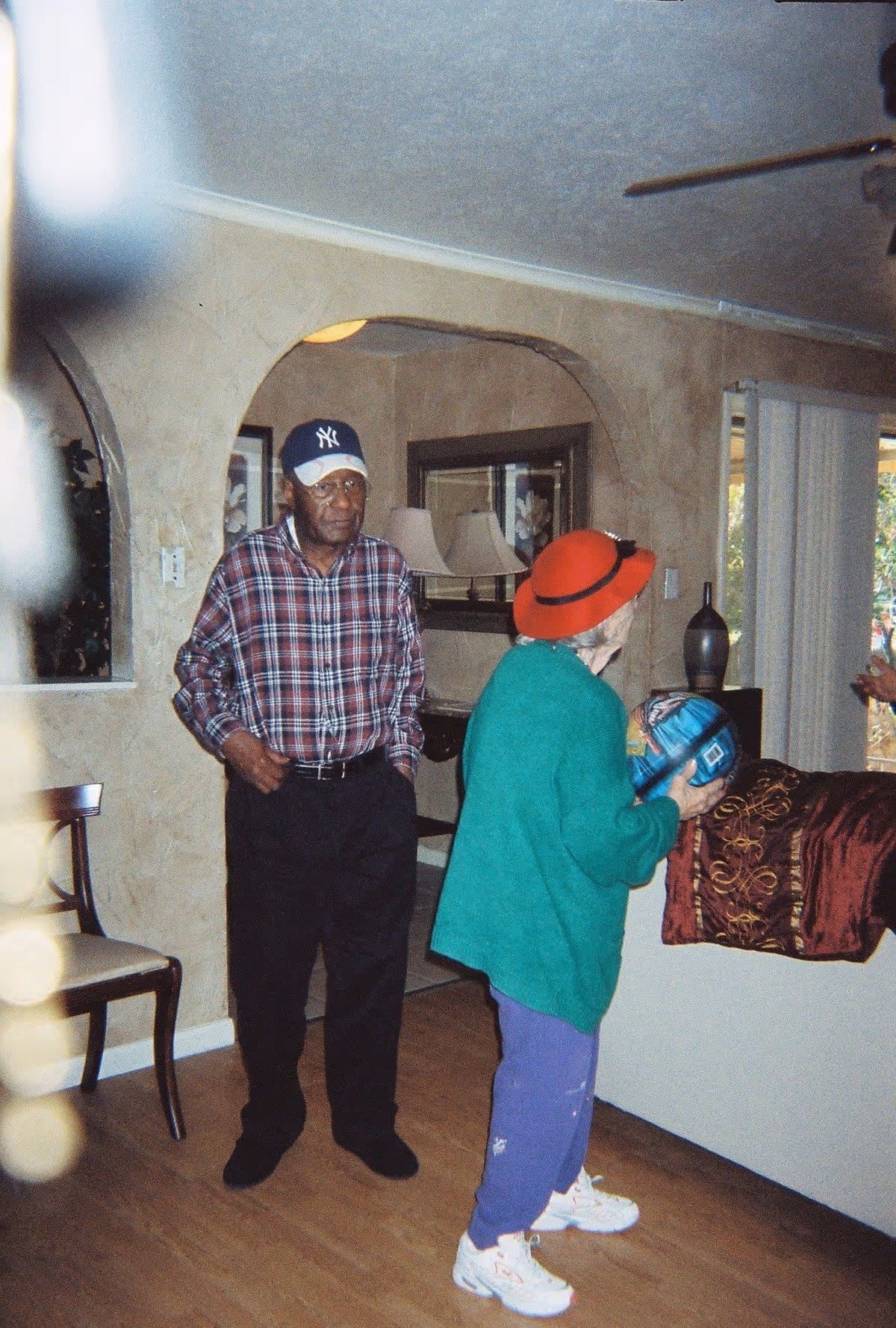 An elderly man wearing a plaid shirt, black pants, and a New York Yankees baseball cap stands with his hands in his pockets inside a living room. An elderly woman wearing a red hat, green sweater, purple pants, and white sneakers is holding a blue ball and facing away from the camera. The room has beige walls, wooden flooring, a chair, a mirror, a lamp, and a window with vertical blinds.
