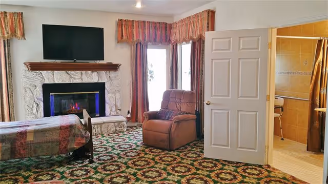A cozy room with a patterned carpet, a bed with a patchwork quilt, a brown recliner chair, and a fireplace with a TV mounted above it. The room has large windows with red and gold curtains. An open door reveals a bathroom with tan tiles and a shower curtain.