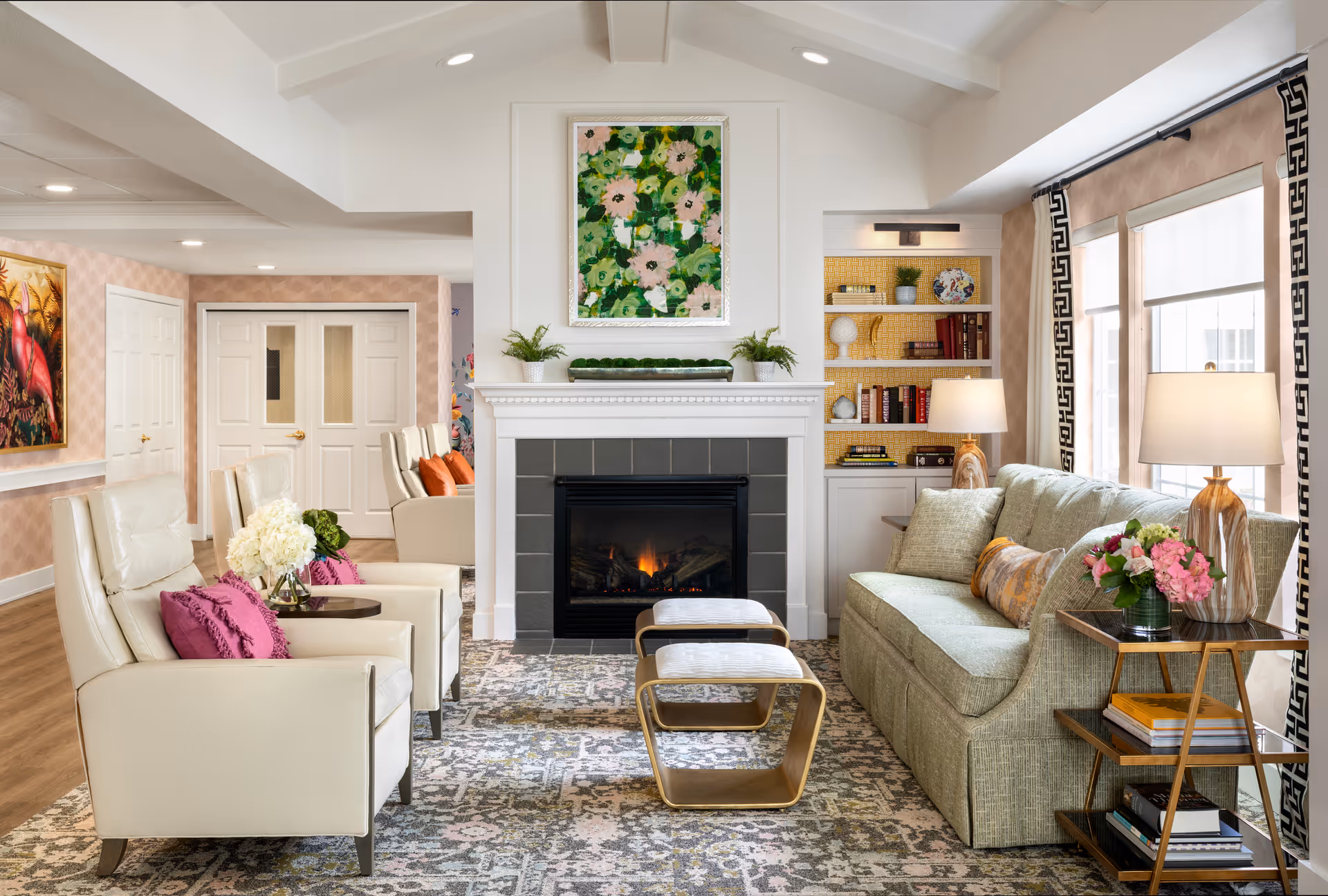 A cozy living room with a fireplace in the center, flanked by built-in shelves with books and decorative items. There is a green sofa with patterned pillows on the right, two cream-colored armchairs with purple cushions on the left, and two small tables in front of the fireplace. The room is well-lit with natural light from large windows on the right and two table lamps. A colorful floral painting hangs above the fireplace.