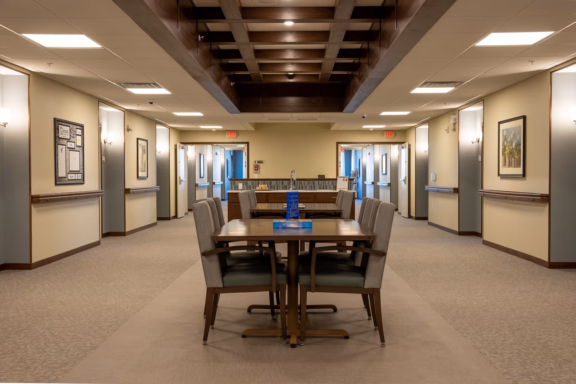 A well-lit hallway in a senior living facility with a wooden table and six upholstered chairs in the center. The hallway has beige walls, framed artwork, and multiple doors leading to other rooms. The ceiling features a wooden grid design with recessed lighting.