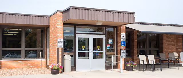 Front entrance of Long Prairie Care Center showing double glass doors with metal frames, brick exterior walls, large windows, a trash can, potted flowers, and several chairs arranged outside under an awning.