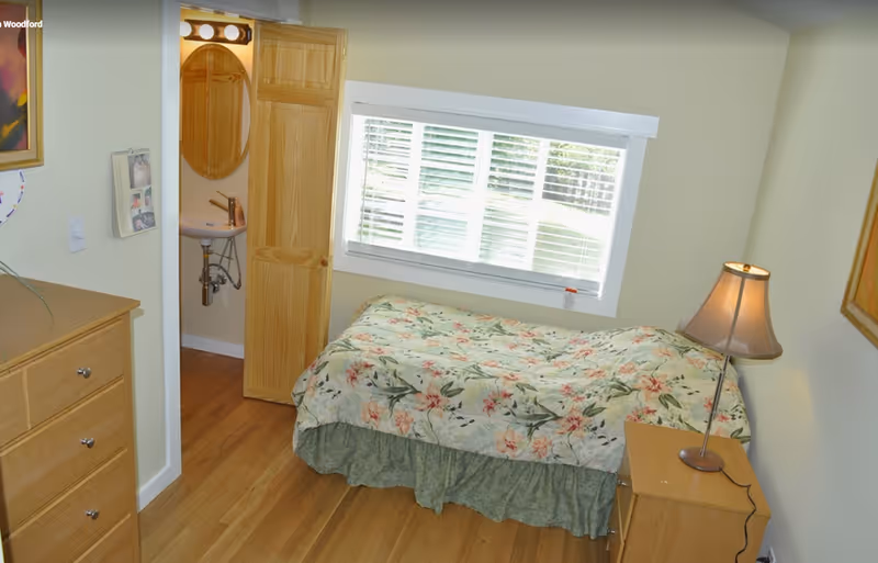 A small bedroom with a single bed covered in a floral bedspread, a wooden nightstand with a lamp, a wooden dresser, and a window with white blinds. There is an open door leading to a small bathroom with a sink and an oval mirror.