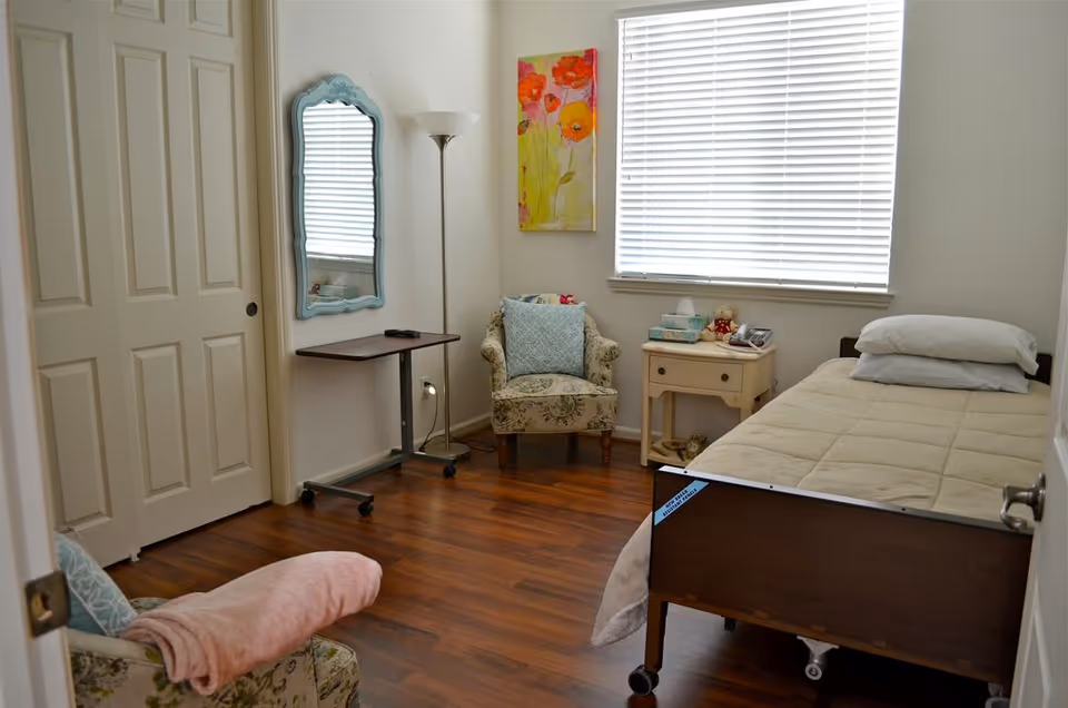 Small furnished bedroom with a single bed, nightstand, two armchairs, a floor lamp, mirror, and a window with blinds.