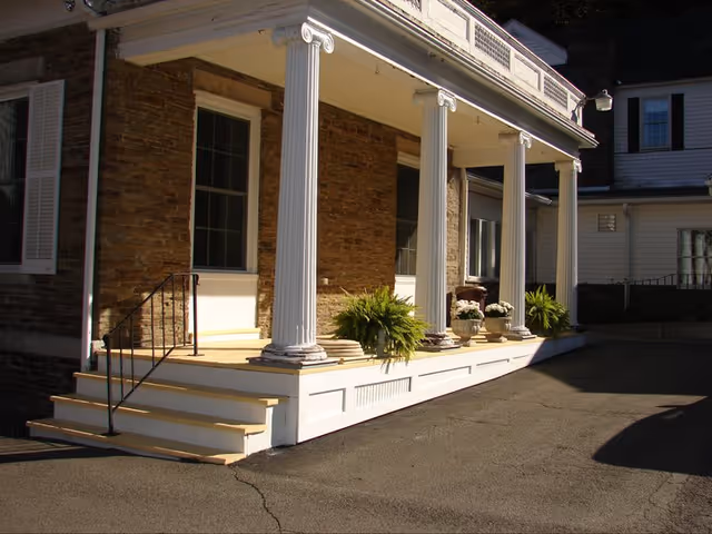 Front porch of a building with white columns, steps leading up to the porch, potted plants, and brick walls under a clear sky.