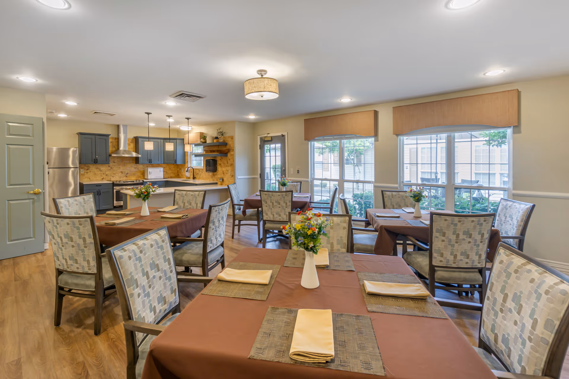 A bright dining room with several tables covered in brown tablecloths, each set with placemats, folded napkins, and a vase of colorful flowers. The room features patterned cushioned chairs, large windows with beige valances letting in natural light, and a kitchen area with blue cabinets, stainless steel appliances, and pendant lighting in the background.