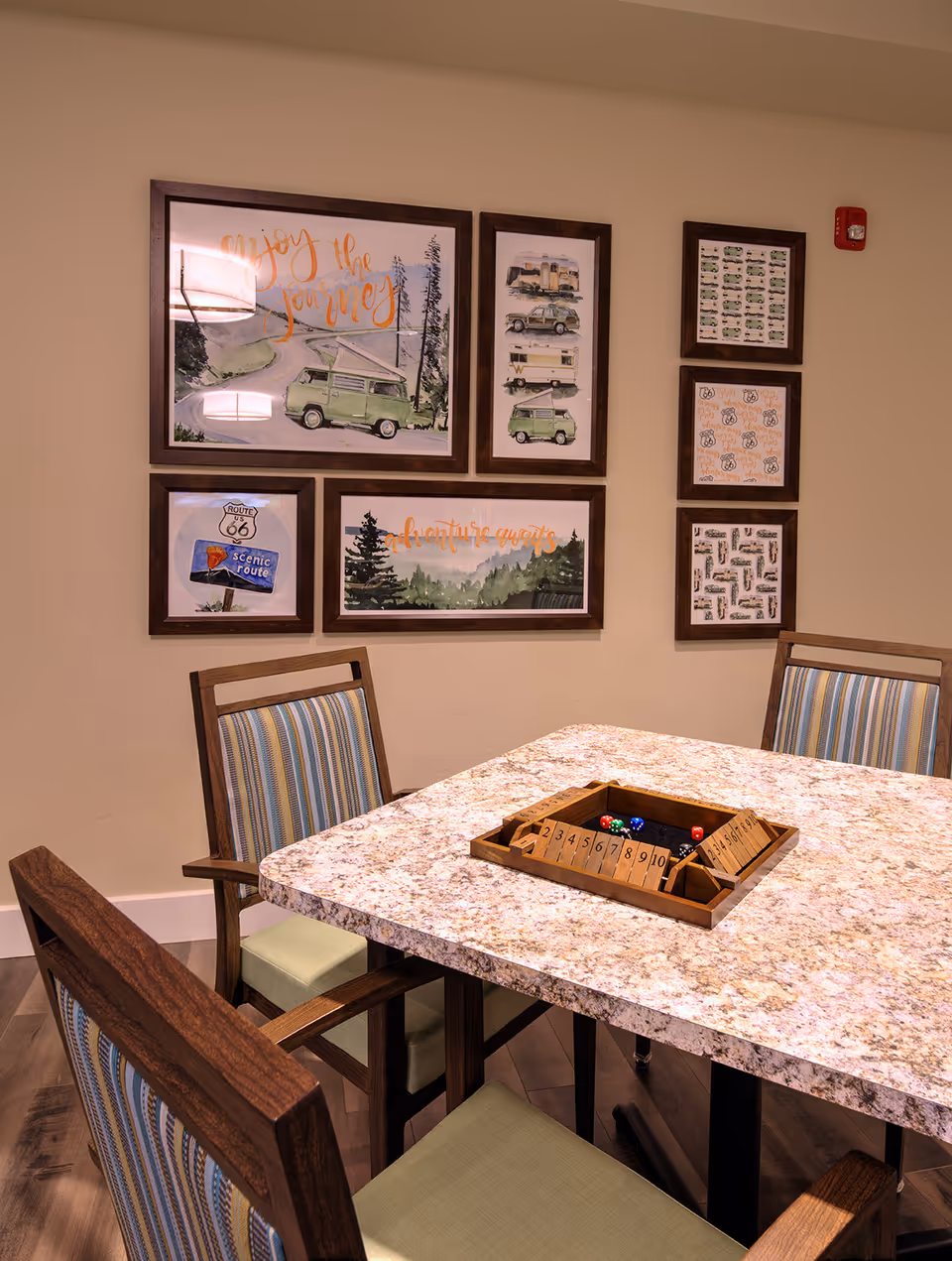 A granite-topped game table surrounded by wooden chairs with striped cushions and framed travel-themed artwork on a beige wall.