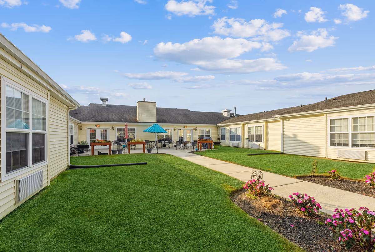 Sunny courtyard with a green lawn, central walkway, patio tables with umbrellas, and surrounding single-story buildings.