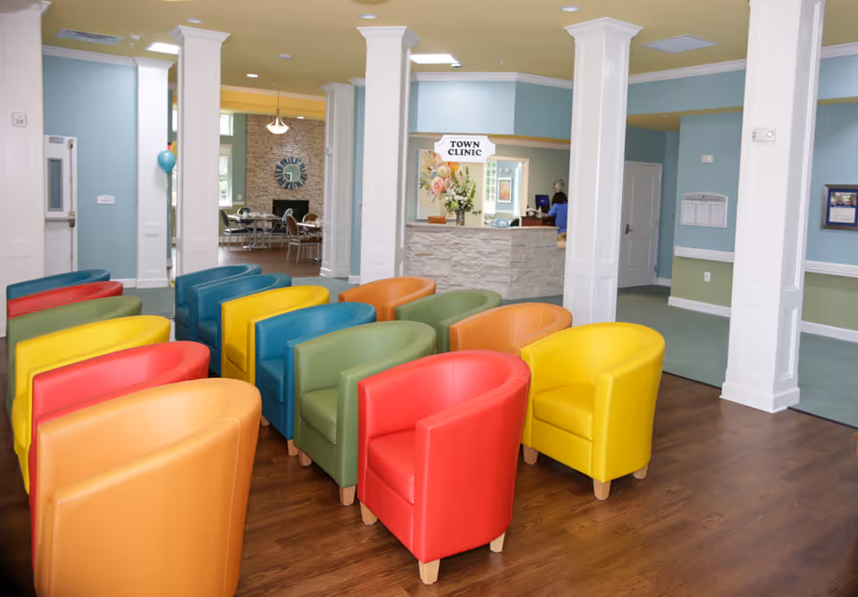 A brightly colored waiting area with multiple rows of armchairs in red, yellow, green, blue, and orange arranged on a wooden floor. In the background, there is a reception desk with a sign that reads 'Town Clinic' and a person working behind the desk. The walls are painted light blue and green, and white columns separate the spaces. A dining area with tables and chairs is visible further back.
