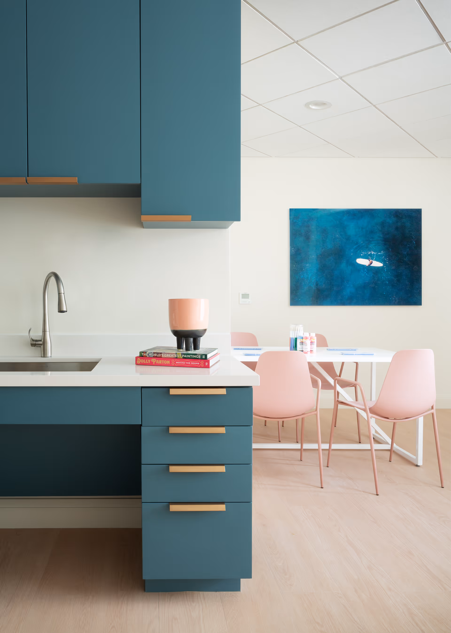 Modern kitchen area with teal cabinets and gold handles, a stainless steel sink with a curved faucet, and a white countertop holding a pink and black cup and two stacked books. In the background, there is a white dining table with four pink chairs and a blue abstract painting on the wall.