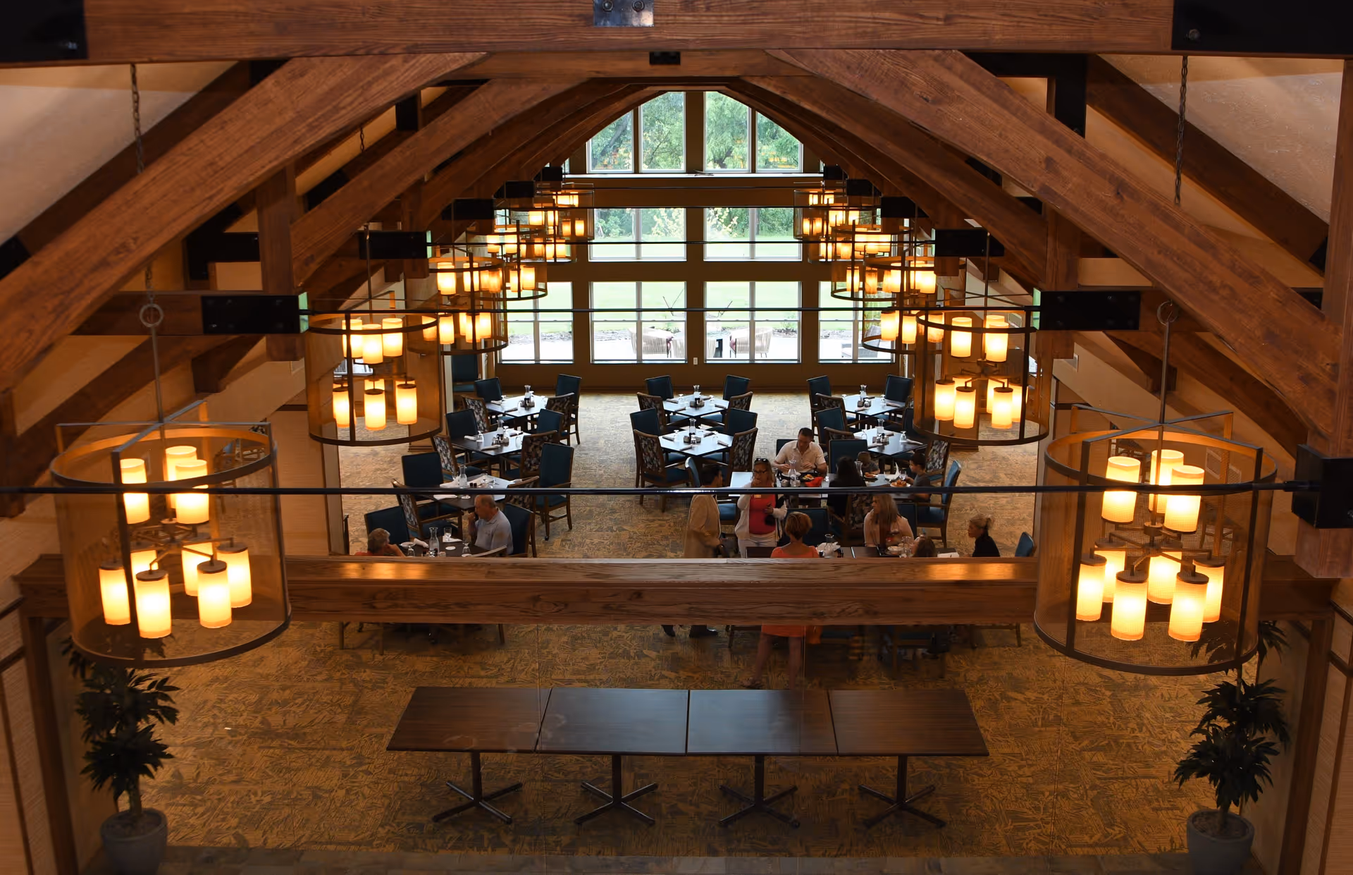 Interior view of a dining room in a retirement community with wooden beams on the ceiling, multiple hanging chandeliers with warm lights, several tables with chairs, and people seated and standing around the tables near large windows showing greenery outside.