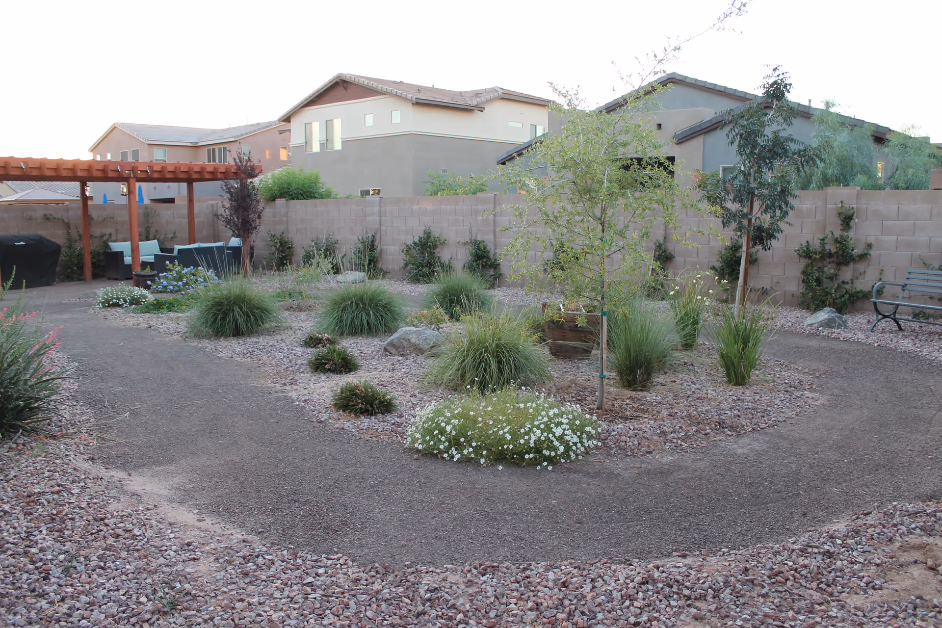 A landscaped outdoor garden area with a gravel pathway winding through various plants and small trees. There is a wooden pergola with outdoor seating and a barbecue grill on the left side. A stone wall encloses the garden, and residential buildings are visible in the background.