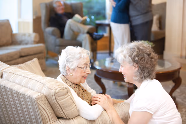 Two elderly women sitting and smiling at each other in a cozy living room with a beige couch and a round wooden coffee table. In the background, a man is seated in an armchair reading a book, and another couple is standing and embracing.