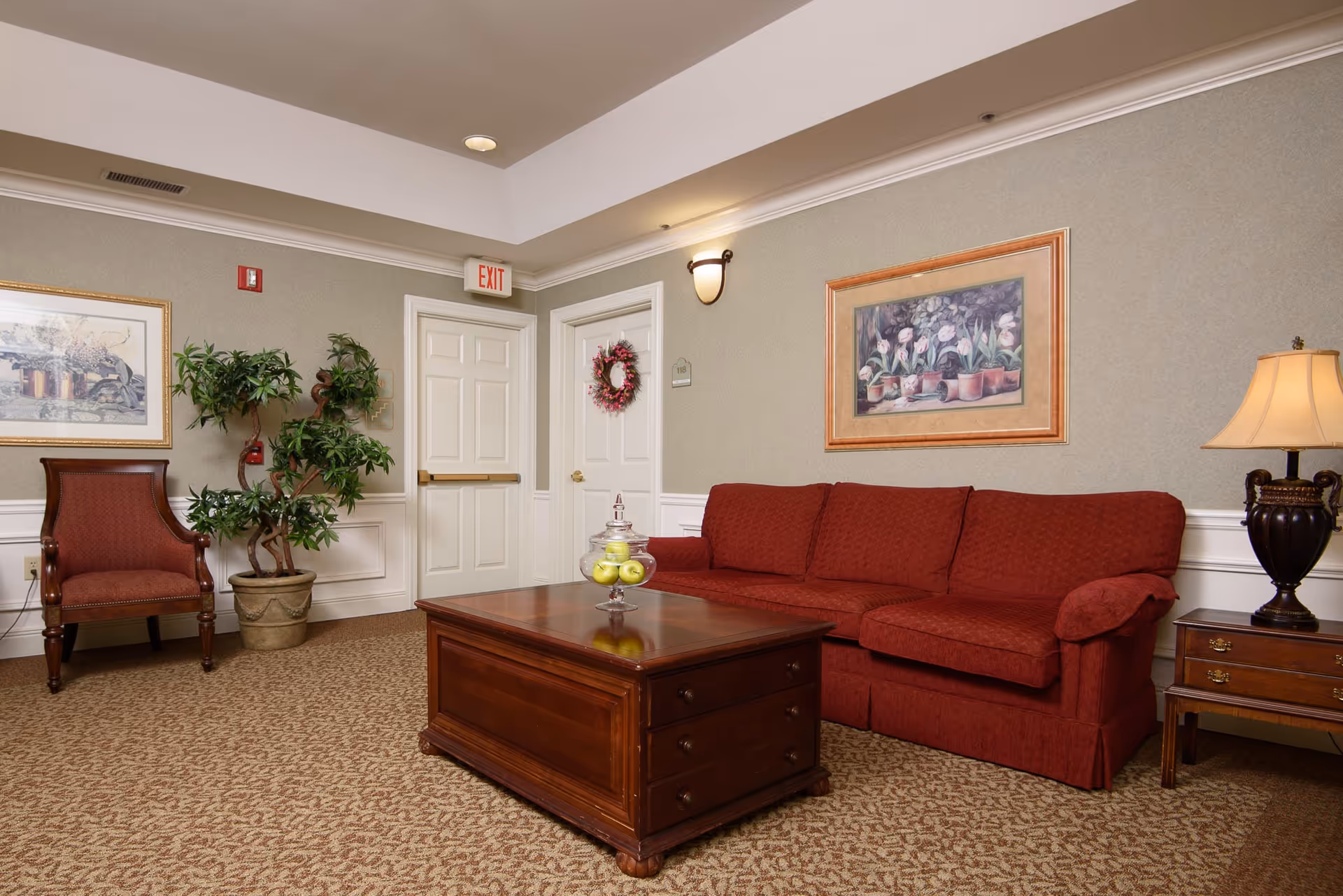 A cozy sitting area in a senior living facility featuring a red upholstered sofa, a wooden coffee table with a glass jar of green apples, a wooden side table with a lamp, a red armchair, a potted plant, framed artwork on the walls, and two white doors, one with a wreath and the other with an exit sign above it.