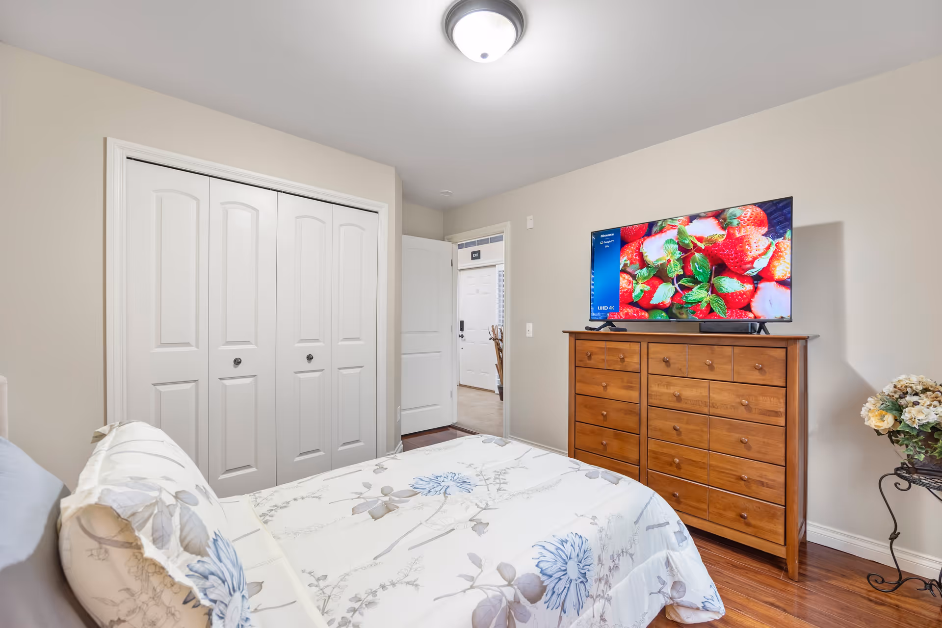 A bedroom with a bed covered in a white floral comforter with blue and gray flowers. There is a wooden dresser with multiple drawers against the wall, topped with a flat-screen TV displaying an image of strawberries and mint leaves. To the left, there is a closed white double-door closet, and an open door leading to another room. A small table with a flower arrangement is visible on the right side of the dresser. The room has light-colored walls and wooden flooring.