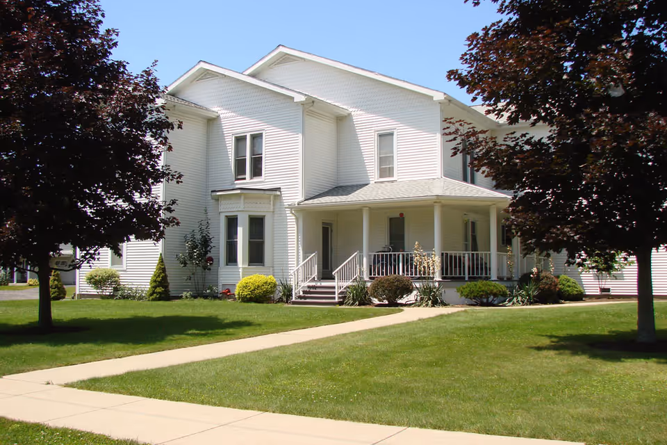 White two-story house with a covered front porch, manicured lawn, sidewalk, and trees.