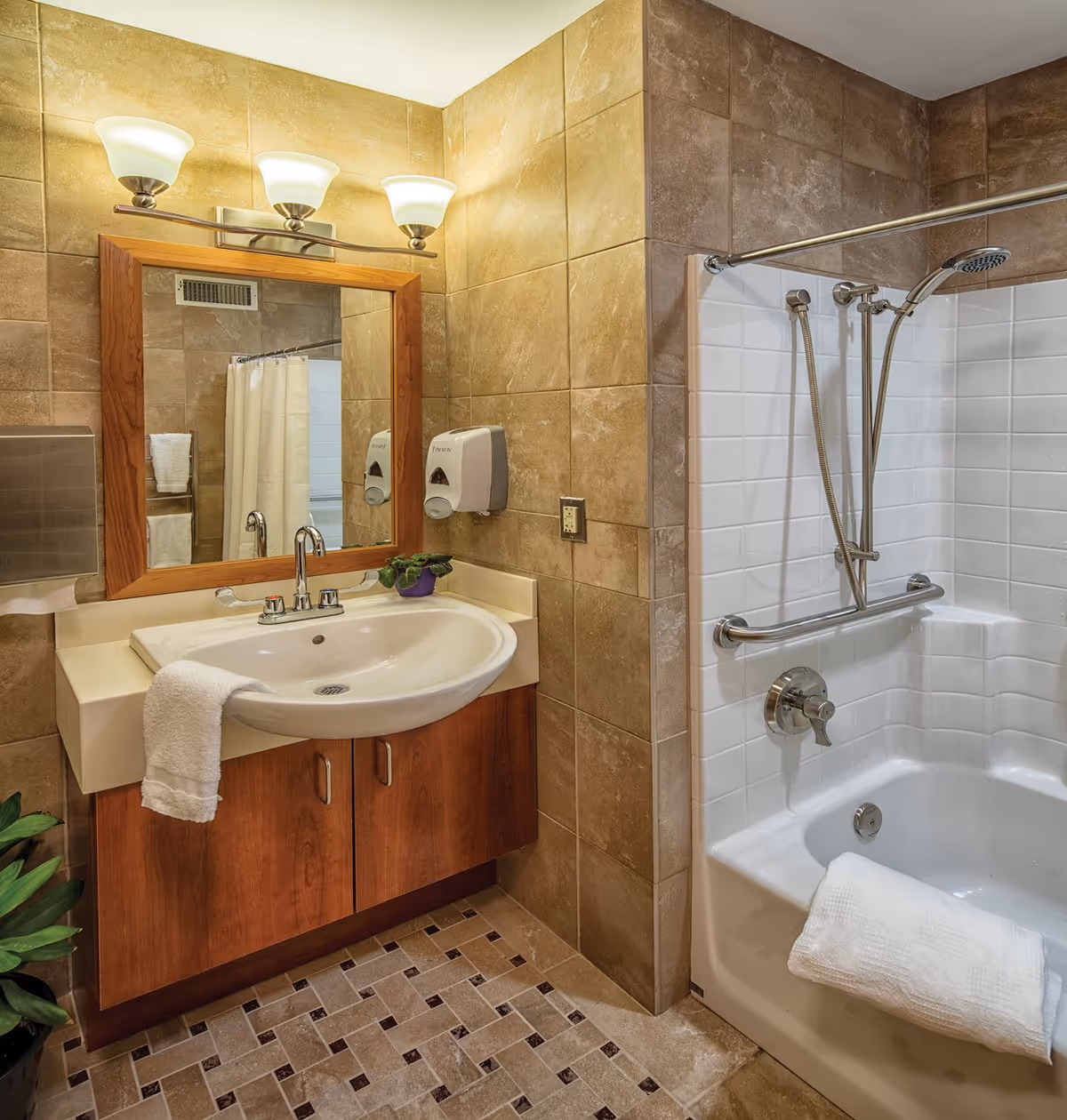 A bathroom with beige tiled walls and floor, featuring a white sink with a wooden cabinet below, a large mirror with a wooden frame above the sink, and three light fixtures mounted above the mirror. To the right is a white bathtub with a showerhead, grab bar, and a white towel draped over the edge. There is also a soap dispenser mounted on the wall next to the sink.