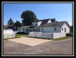 Exterior view of a single-story white residential building with a white picket fence surrounding a small yard, a concrete driveway, and a large tree in the background under a clear blue sky.