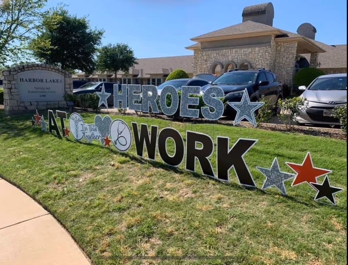 Outdoor view of Harbor Lakes Nursing and Rehabilitation Center with a large decorative sign on the grass that reads 'HEROES AT WORK' with stars and symbols including a heart and stethoscope, and a stone sign displaying the facility name.