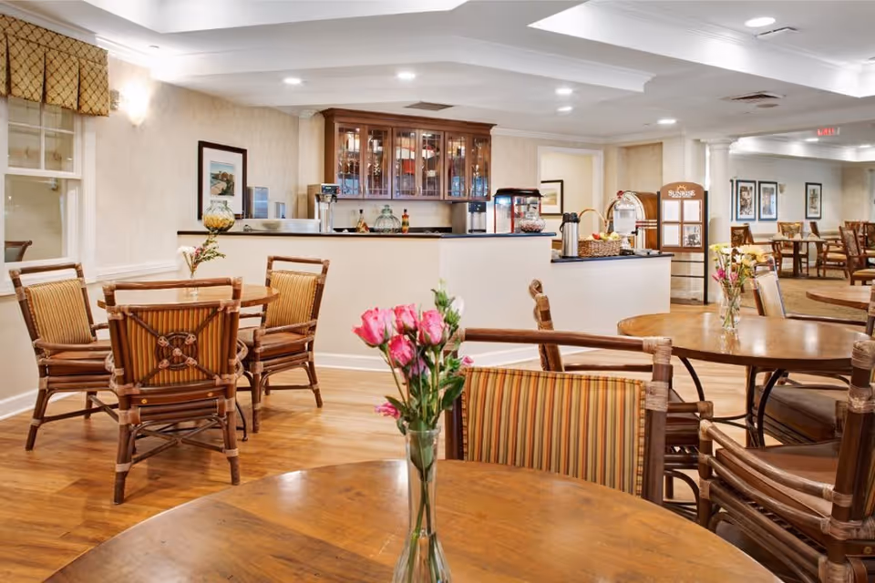 A bright and inviting dining area in a senior living facility with wooden tables and chairs featuring striped cushions. Fresh flowers in vases are placed on the tables. In the background, there is a counter with a coffee machine, popcorn machine, and glass cabinets. The room has warm lighting and a polished wooden floor.