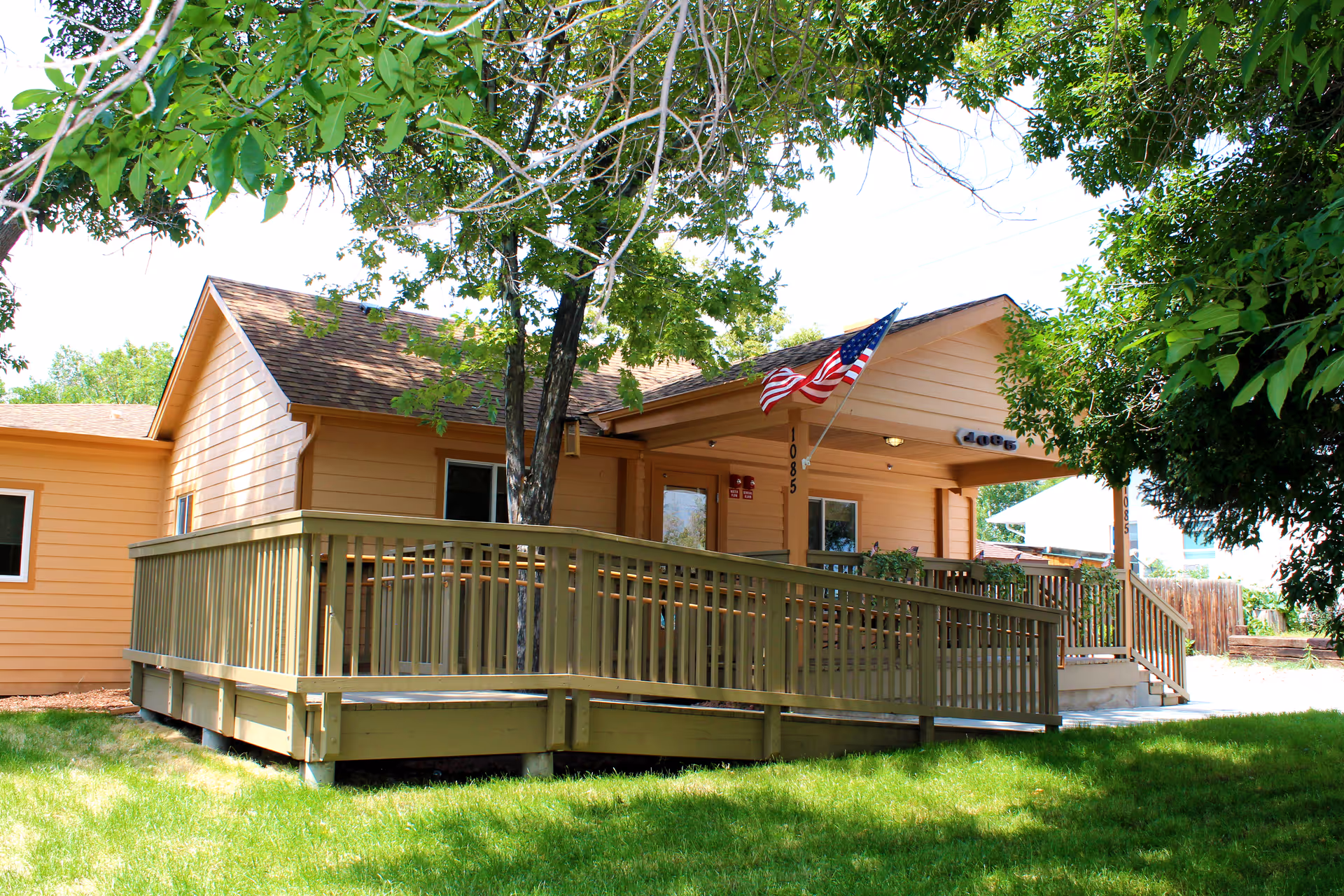 Exterior view of a single-story building with light brown siding and a wooden ramp leading to the entrance. An American flag is mounted near the door, and there are trees and green grass surrounding the building.