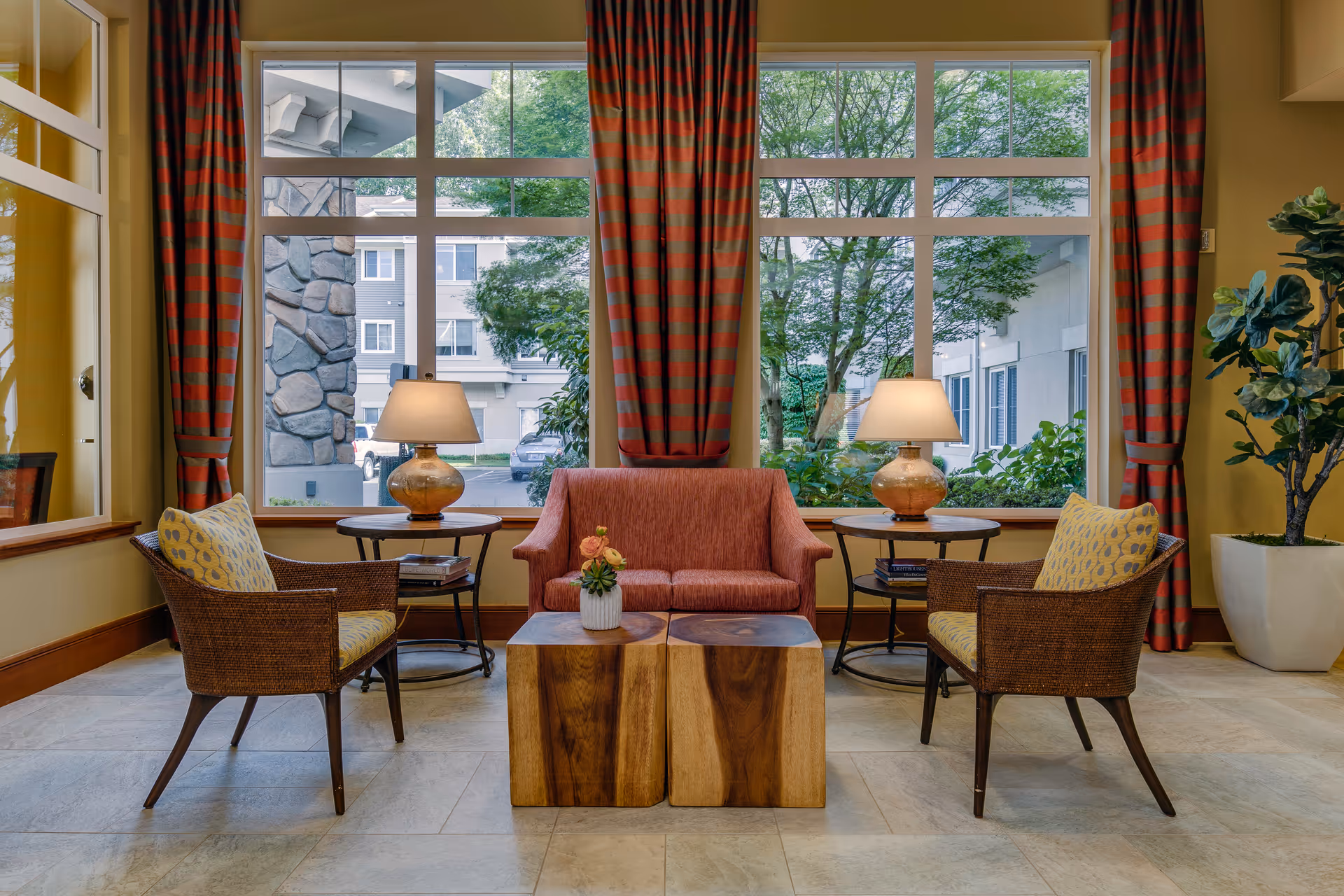 Seating area with a loveseat, two wicker armchairs, wooden coffee tables and lamps in front of large windows showing trees and a building outside.