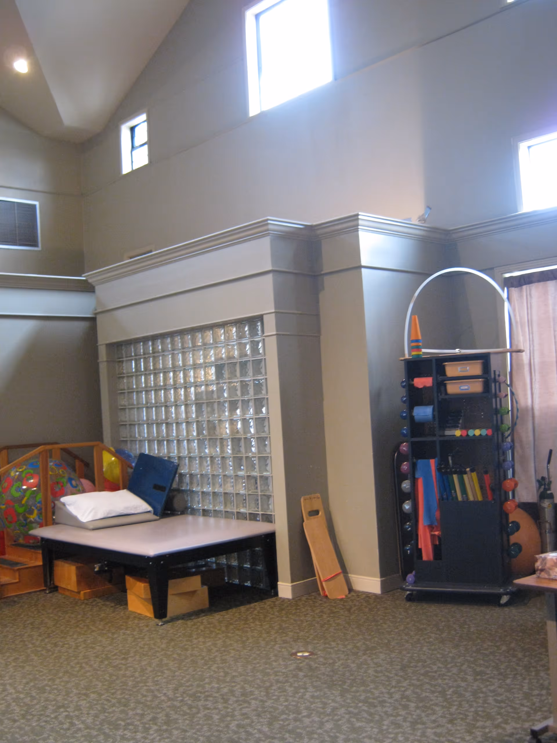 High-ceilinged interior room with exercise/therapy equipment, a padded treatment table and shelving by a glass block wall.