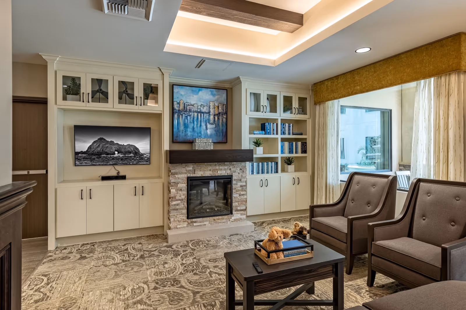 A cozy living room with two brown armchairs, a dark wooden coffee table with a tray holding two teddy bears and a book, a fireplace with a stone surround, built-in white cabinets with glass doors, shelves filled with books and decorative plants, a large window with curtains, and a ceiling with recessed lighting.