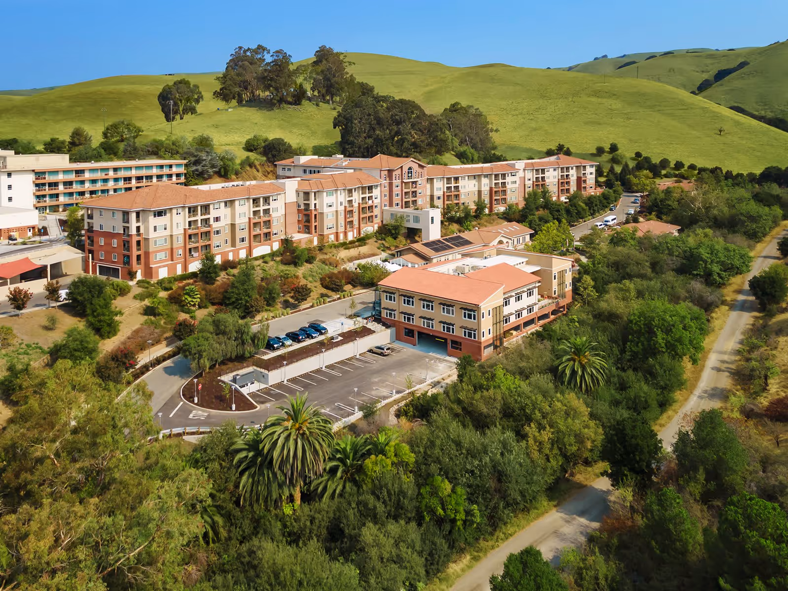 Aerial view of the Masonic Homes of California facility showing multiple multi-story residential buildings with red-tiled roofs surrounded by greenery and hills under a clear blue sky. There is a parking lot with several cars and a road running alongside the property.