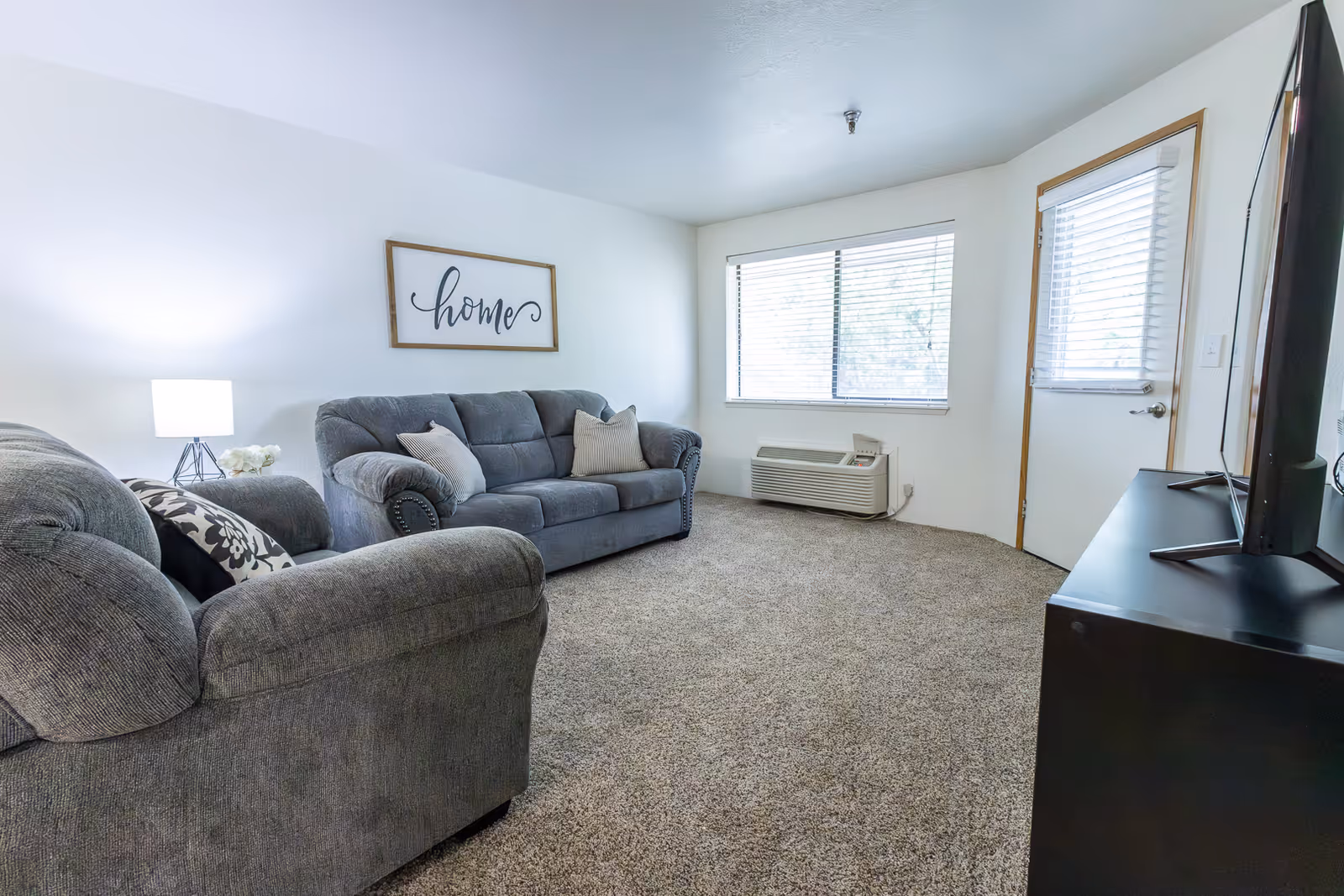 A cozy living room with two gray upholstered sofas, a small side table with a lamp and flowers, a large window with blinds, a door with a window, a wall-mounted air conditioning unit, and a TV on a black stand. A framed sign on the wall reads 'home'.