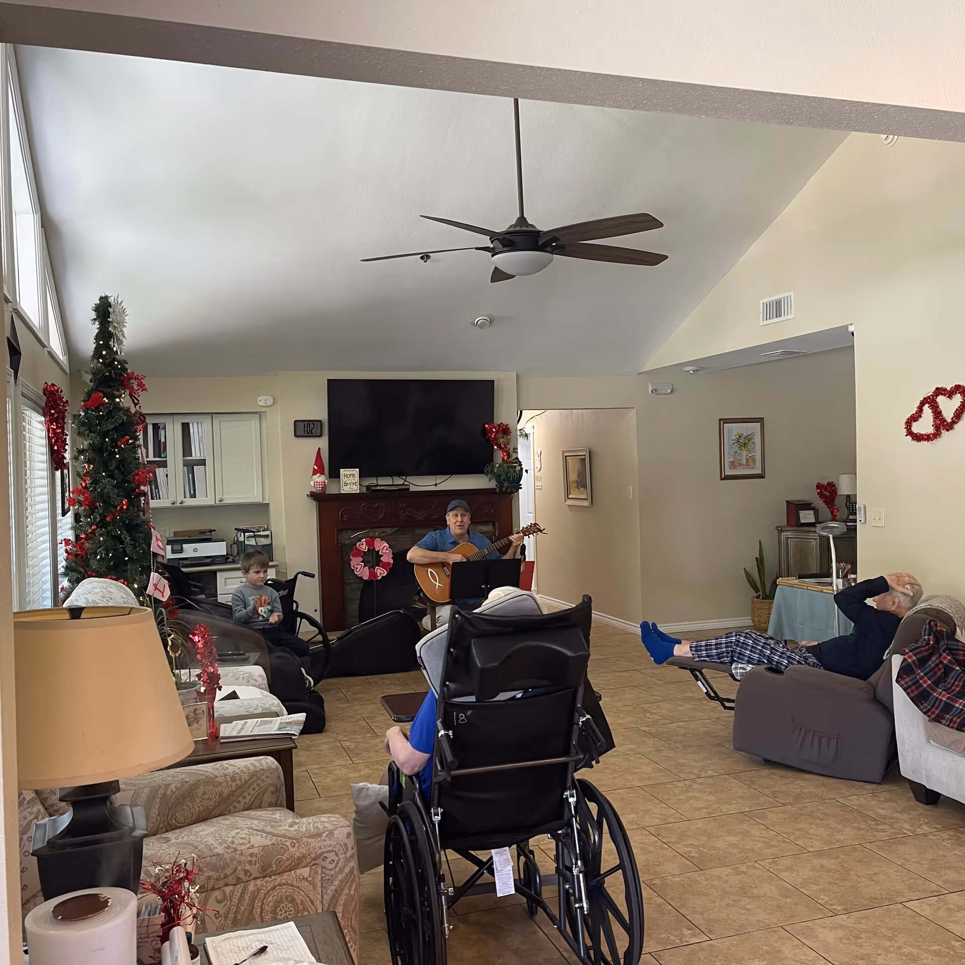 A decorated communal living room with seated residents, a person playing guitar by a TV, and a Christmas tree.