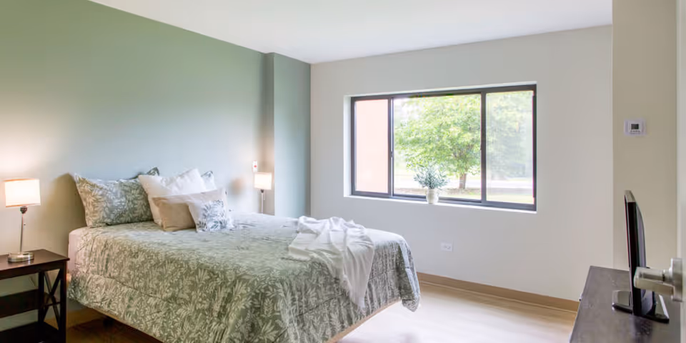 A bright and clean bedroom with a bed covered in green and white floral bedding, two bedside tables with lamps, a large window showing greenery outside, and a TV on a stand opposite the bed.