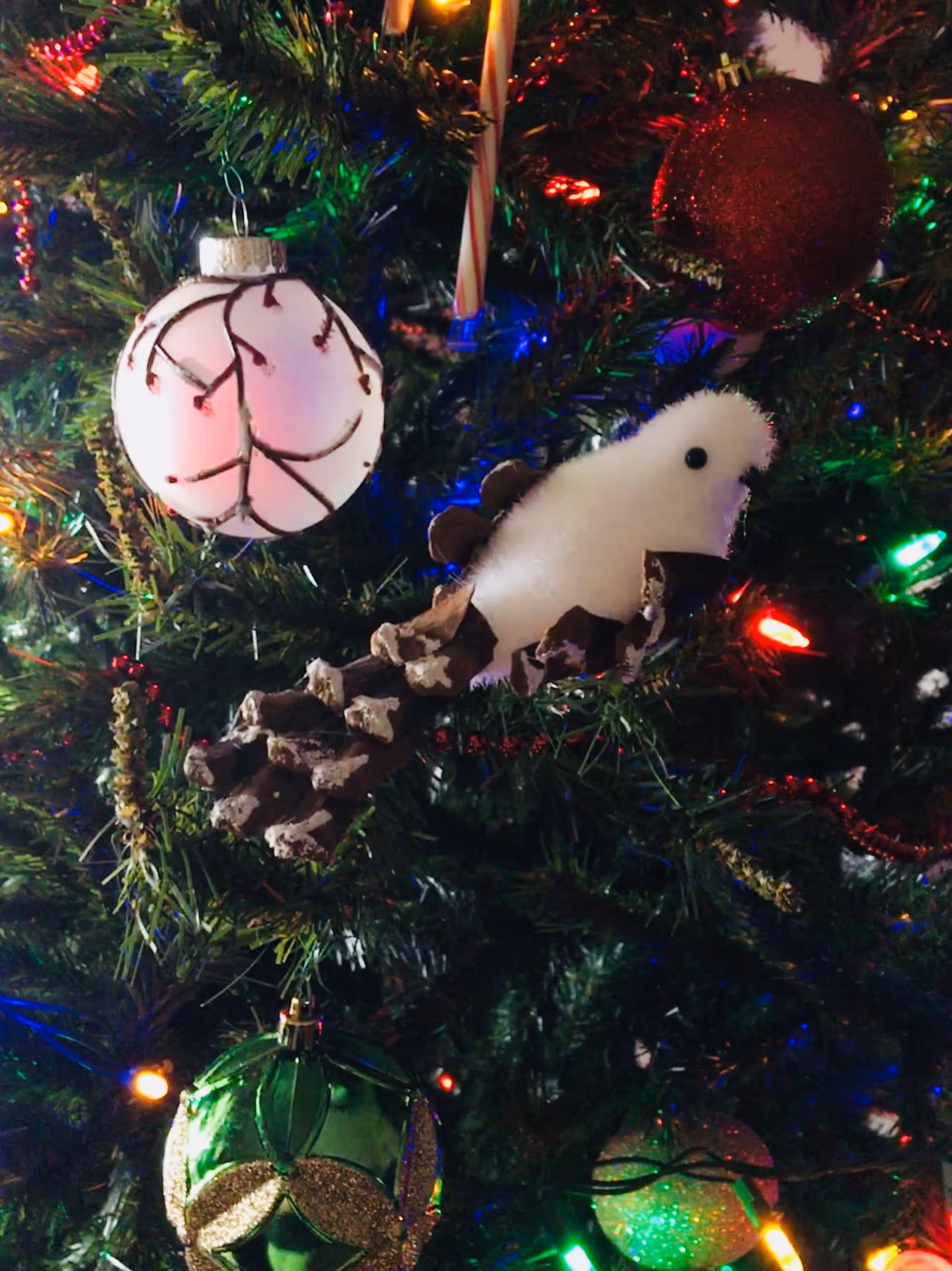 Close-up of a decorated Christmas tree with colorful lights and ornaments, including a white ornament with brown branch-like patterns, a red glittery ball, a green ornament with gold glitter, a candy cane, and a bird-shaped ornament made from a pinecone and white fluffy material.