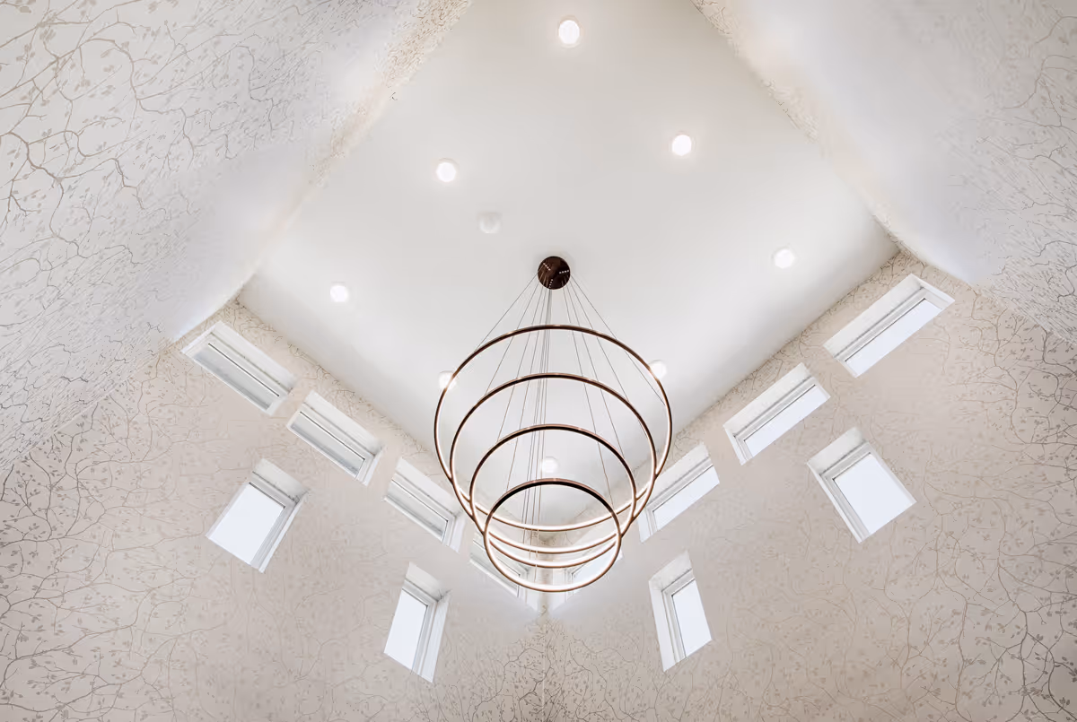 View looking up at a modern ceiling with multiple recessed lights and a large circular chandelier with concentric rings hanging in the center. The walls have a subtle floral pattern and feature several rectangular windows near the ceiling.