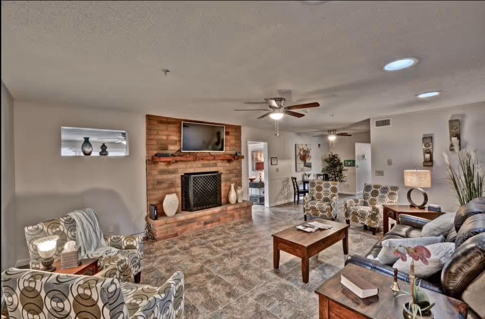 Communal living room with a brick fireplace and mounted TV, multiple armchairs and sofas arranged around a coffee table and ceiling fans overhead.