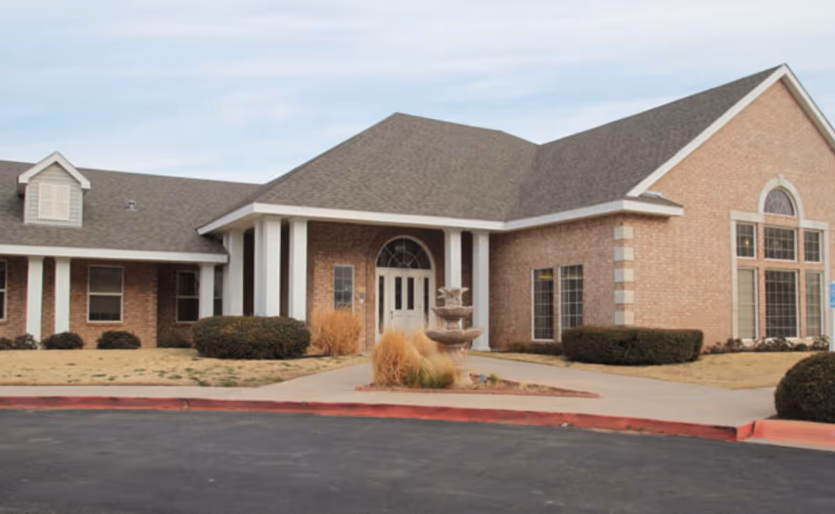 Exterior view of a single-story brick building with a gray shingled roof, white columns at the entrance, large windows, and a small fountain surrounded by landscaping in front of the entrance.