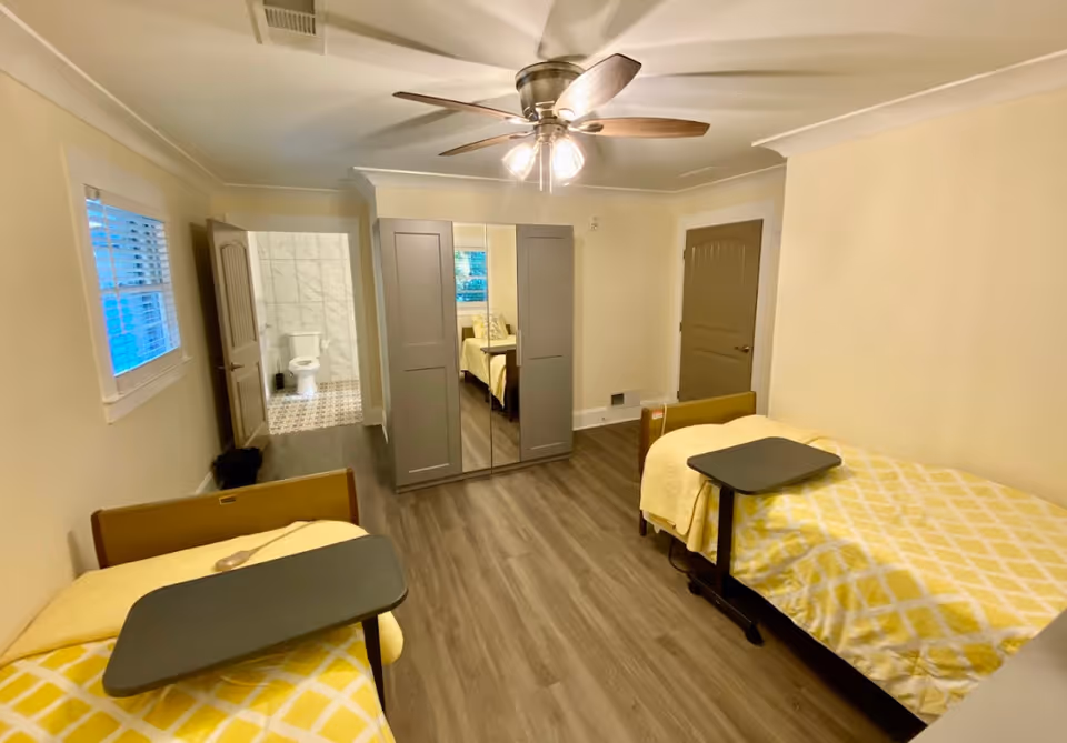 A senior living bedroom with two single beds covered in yellow and white patterned blankets. Each bed has a movable overbed table. The room has wood flooring, a ceiling fan with lights, a large wardrobe with mirrored doors, and two closed doors. One door leads to a bathroom with a visible toilet and tiled walls and floor. A window with blinds is on the left wall.