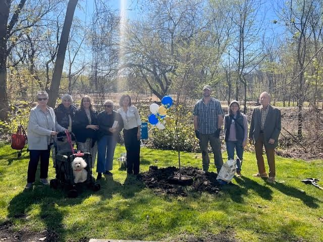 A group of seven people standing outdoors on a grassy area around a newly planted tree decorated with blue and white balloons. There is a small white dog in a stroller, and the background shows leafless trees and a clear sky.