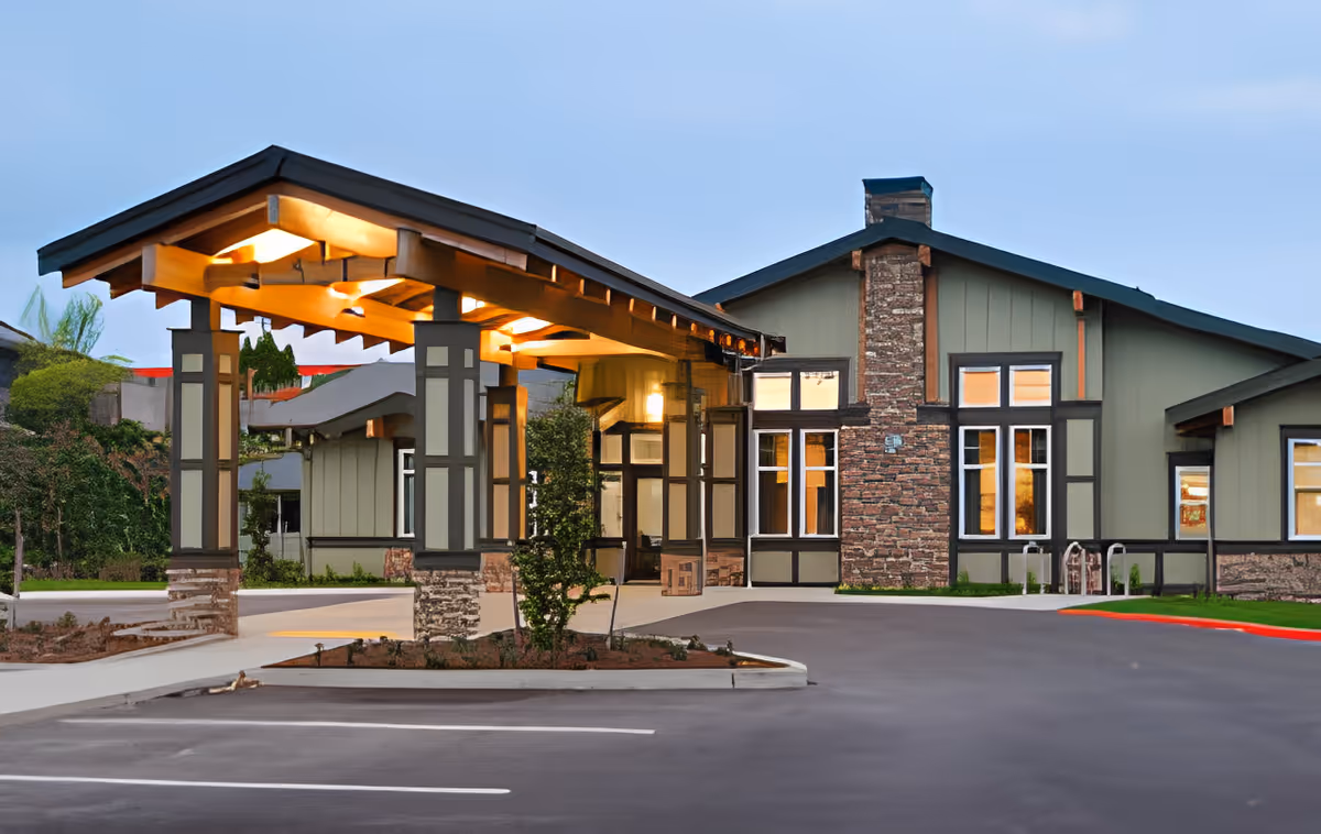 Exterior view of The Arbor at Bremerton senior living facility entrance with a covered drop-off area, stone and wood architectural details, and a paved parking lot in front.