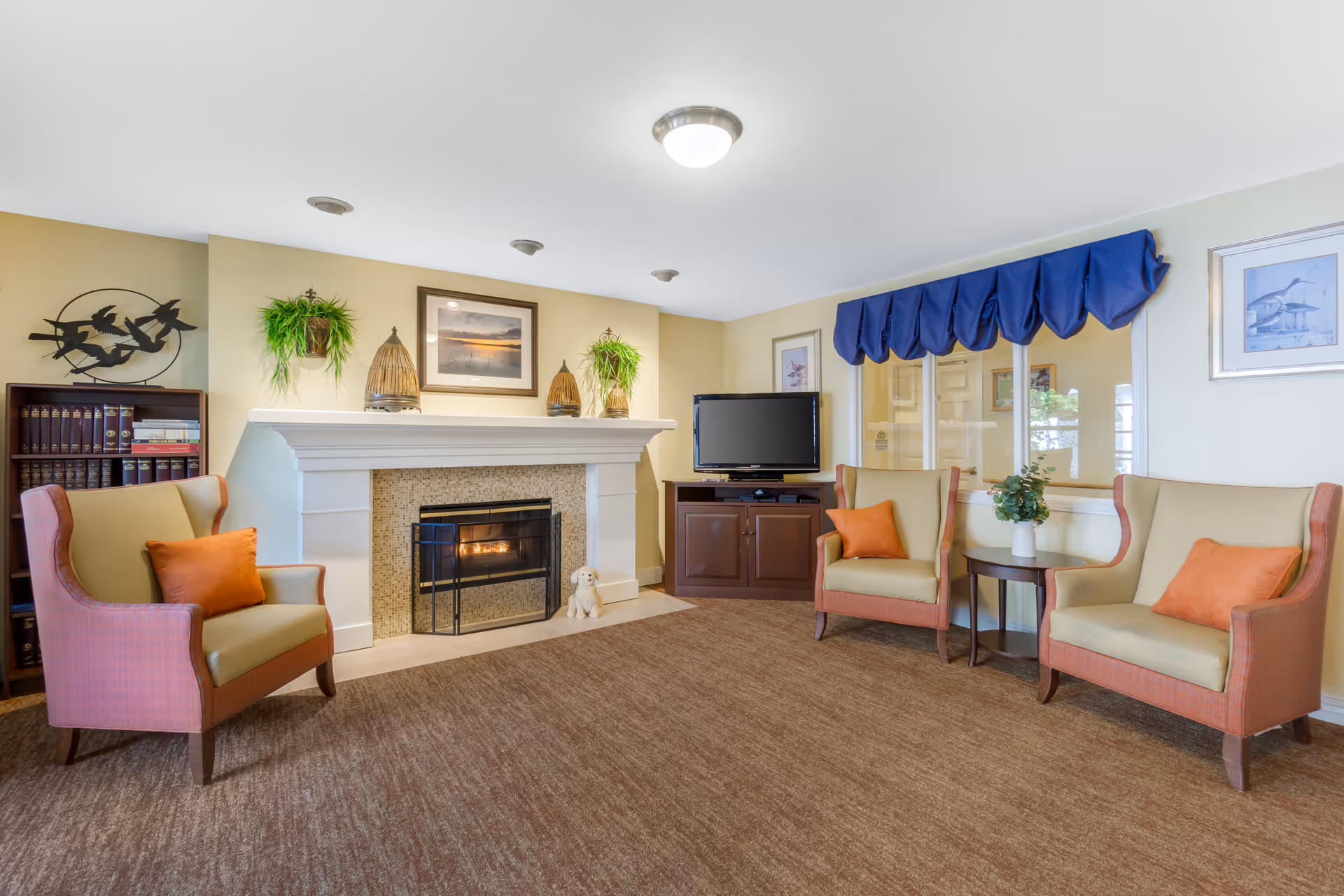 A cozy living room area with three cushioned armchairs each with an orange pillow, a fireplace with a fire burning, a TV on a wooden cabinet, a bookshelf with books, wall art, and two hanging plants. The room has beige walls, a brown carpet, and a window with a blue valance.
