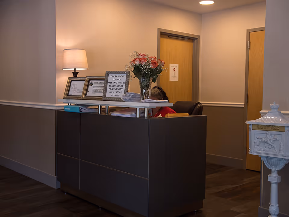 Reception desk in a facility lobby with a lamp, framed notices, a vase of flowers, and a staff member seated behind the desk.