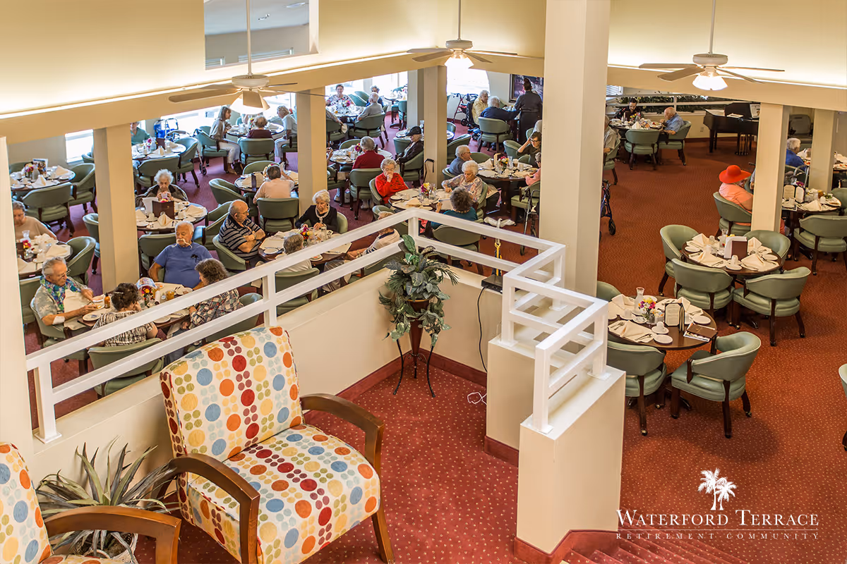 Interior view of a dining room in a retirement community with elderly residents seated at round tables enjoying meals. The room has red carpeting, green cushioned chairs, and ceiling fans. A colorful patterned armchair is visible in the foreground near a white railing. The atmosphere is bright and social.