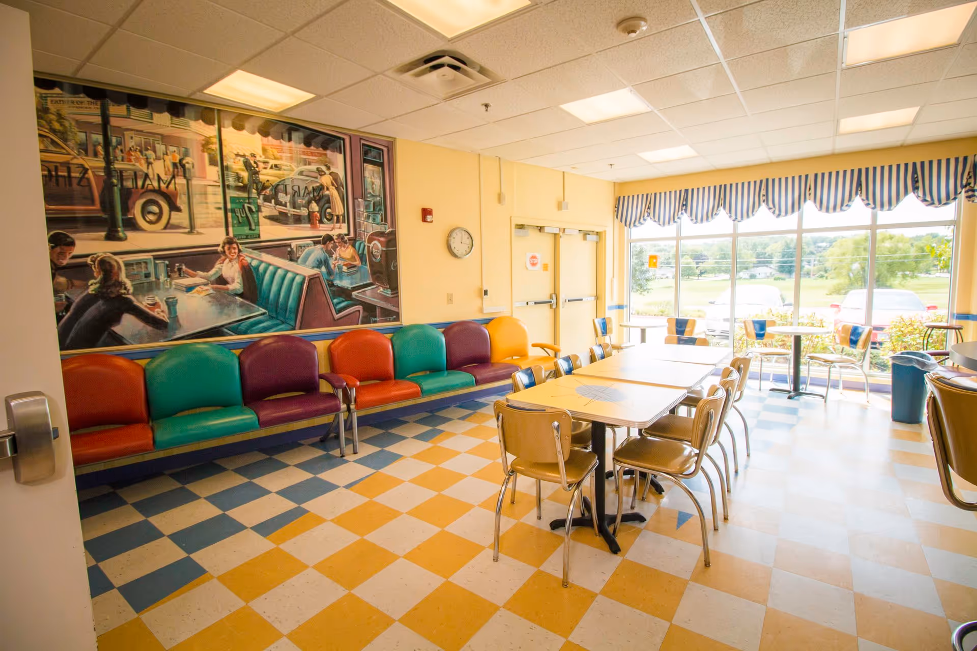 A brightly lit dining area with checkered yellow and blue flooring, colorful chairs lined up against the wall, several tables with chairs, large windows with striped valances letting in natural light, and a large mural on the wall depicting a vintage diner scene.