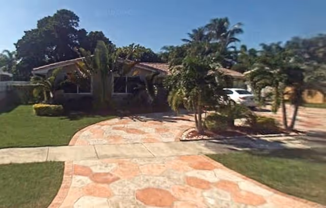 Front exterior view of a single-story building with a tiled roof, surrounded by palm trees and greenery, with a patterned stone driveway and a white car parked on the right side.