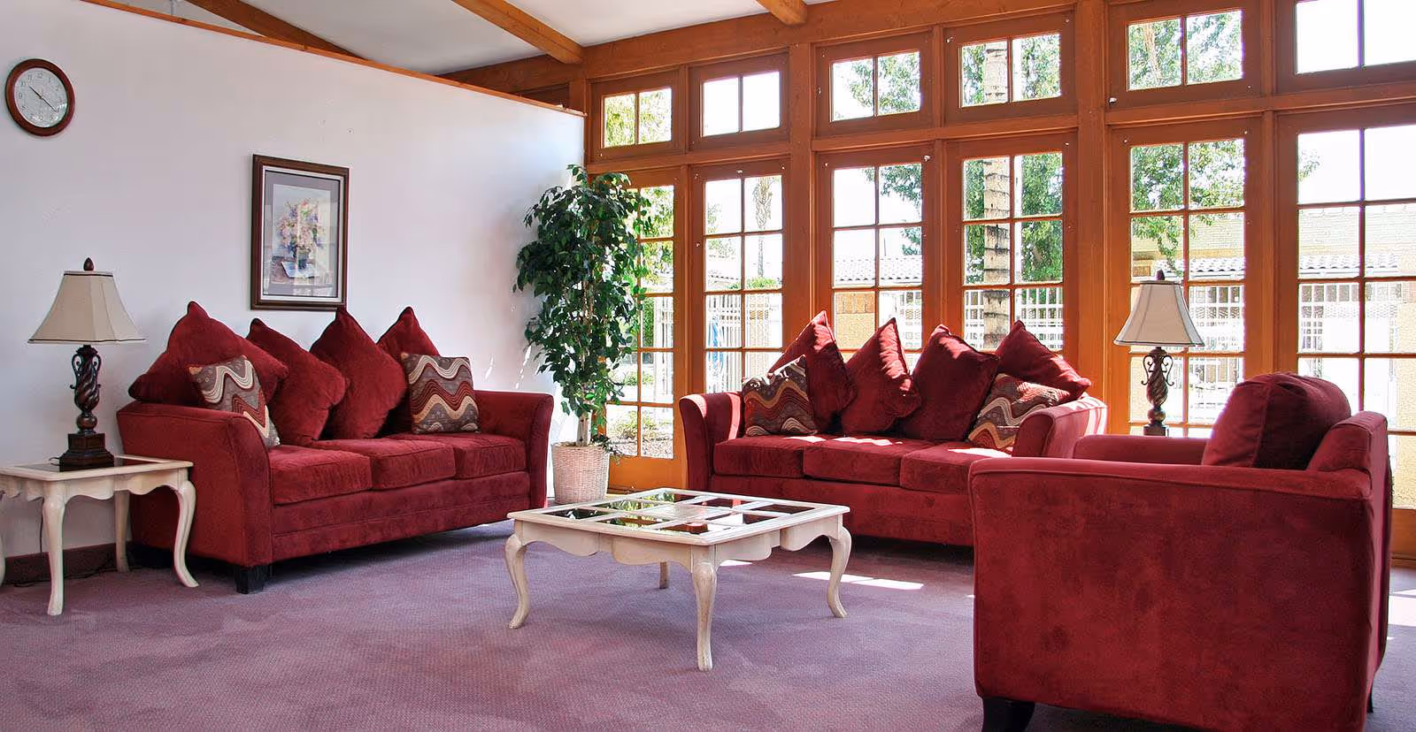 Sunlit living room with red sofas and an armchair arranged around a coffee table in front of large wood-framed windows.