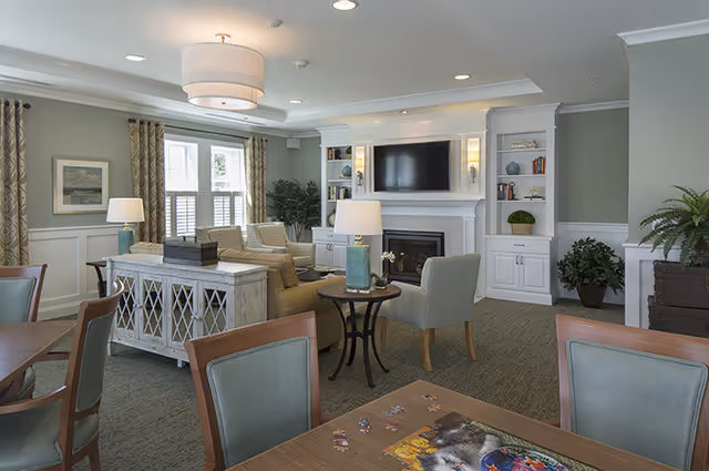 A cozy senior living common area with comfortable armchairs and sofas arranged around a fireplace with a mounted flat-screen TV above it. The room has built-in white shelves with decorative items, a large window with curtains, and several table lamps providing warm lighting. A wooden table with chairs is in the foreground, with a puzzle and a book on it.