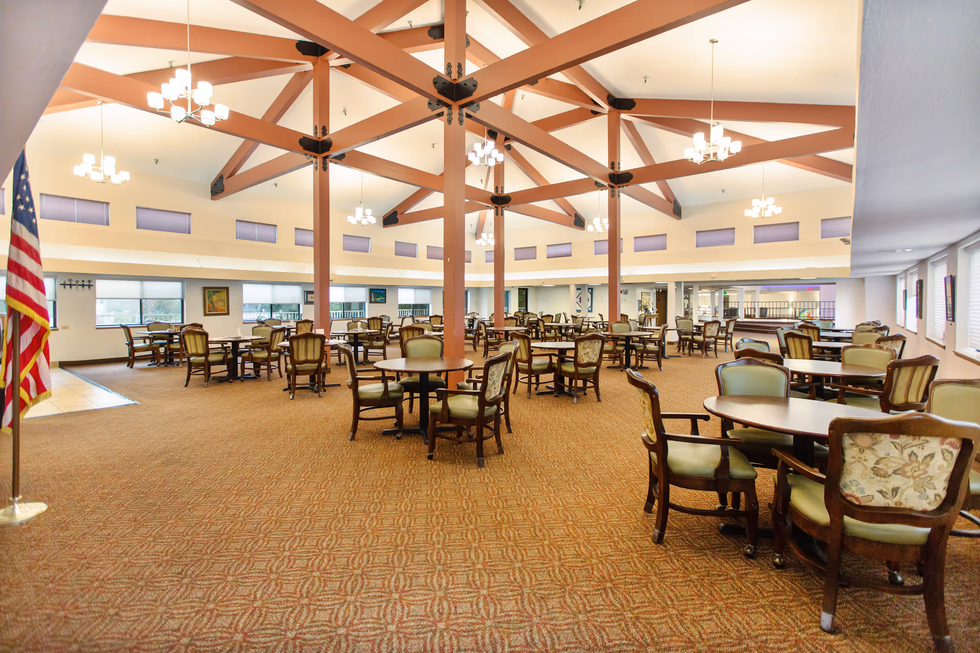 A spacious dining room with multiple round tables and chairs arranged neatly. The room features a high ceiling with exposed wooden beams painted in a reddish-brown color and several modern chandeliers hanging from the ceiling. Large windows line the walls, allowing natural light to fill the space. An American flag is positioned on the left side near the entrance.