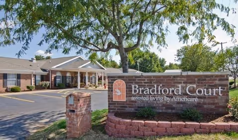 Exterior view of Bradford Court assisted living facility showing a brick sign with the facility name and a building with a covered entrance surrounded by trees and a parking area.