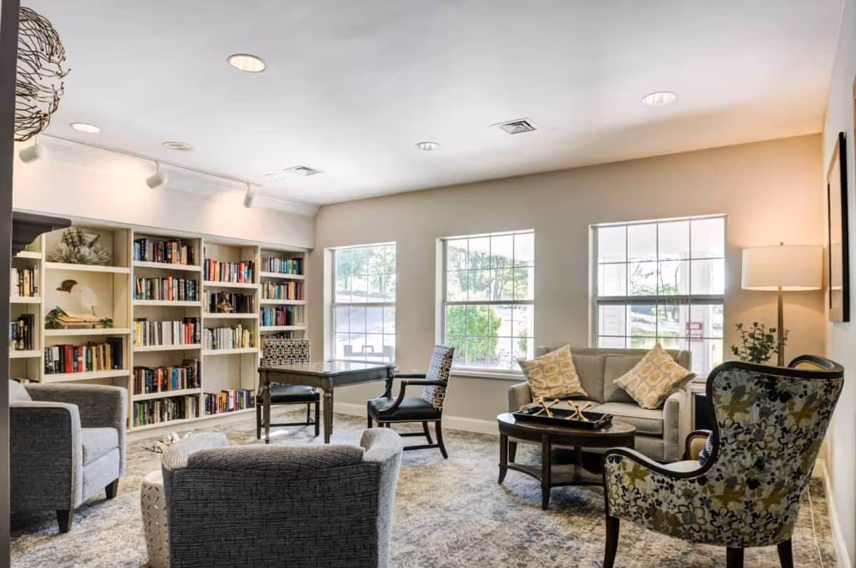 A bright and cozy living room area with large windows letting in natural light. The room features a bookshelf filled with books, a small table with two chairs, a sofa with patterned cushions, a round coffee table, and several upholstered armchairs. A floor lamp and decorative plants add to the inviting atmosphere.