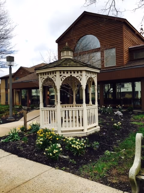 A white wooden gazebo with decorative arches situated in a garden area with blooming yellow and white flowers. Behind the gazebo is a brown building with large windows and a peaked roof. A concrete pathway curves around the garden, and a bench is partially visible on the right side of the image.