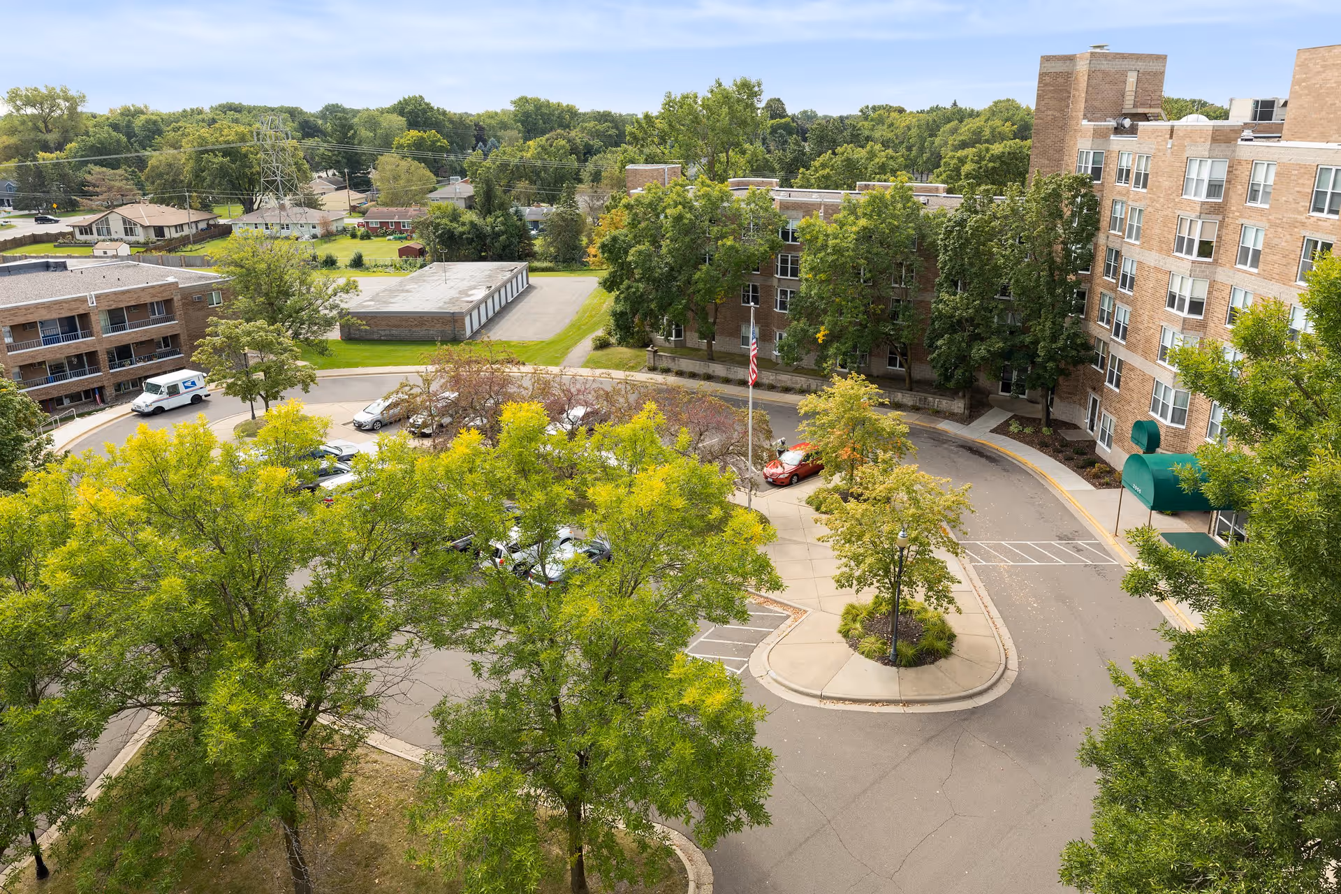 Aerial view of RosePointe Senior Living facility showing a circular driveway with parked cars, surrounded by trees and multi-story brick buildings. The area is lush with greenery and there is an American flag in the center of the driveway.
